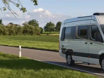 Malibu Genius Campervan auf einer kurvigen Straße mit grünen Feldern und Wasser im Hintergrund